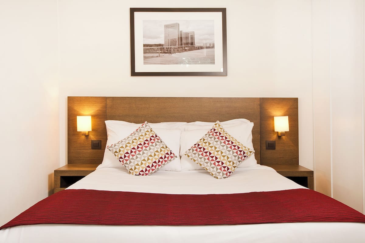Una cama bien hecha con una colcha roja, dos almohadas estampadas y dos lámparas fijadas a la pared.