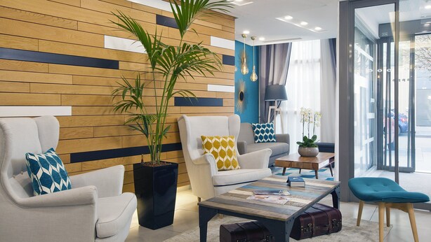 A modern hotel lobby with a wooden accent wall, a large potted plant, a white sofa with a patterned pillow, a coffee table with magazines, and a blue armchair. - Residhome Asnières (Asnieres-sur-Seine)