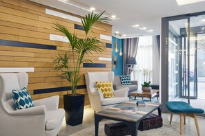 A modern hotel lobby with a wooden accent wall, a large potted plant, a white sofa with a patterned pillow, a coffee table with magazines, and a blue armchair.
