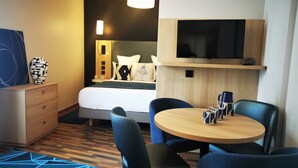 A hotel room with a bed, a TV mounted above it, a wooden dresser, a round table with chairs, and a patterned rug. - Residhome Asnières (Asnieres-sur-Seine)