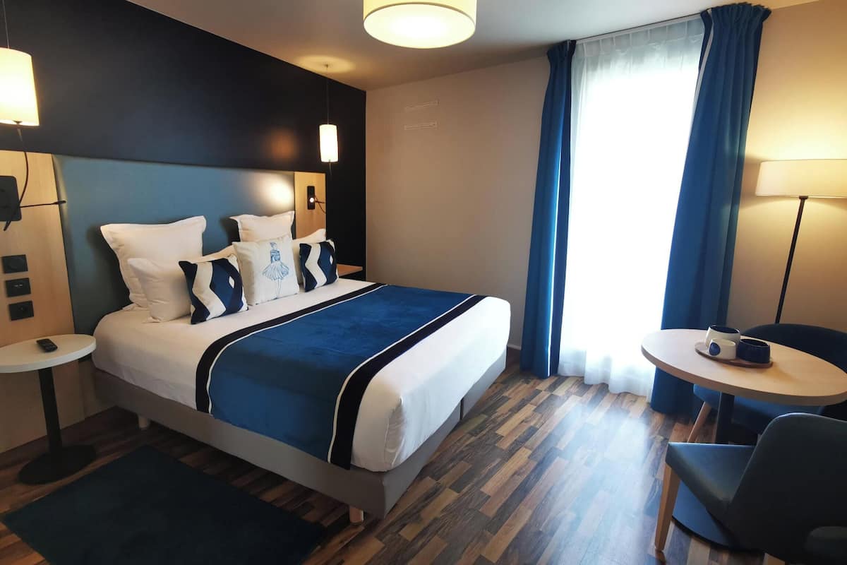 Een hotelkamer met een groot bed, twee nachtkastjes, een ronde tafel met stoelen, een raam met gordijnen en een houten vloer.