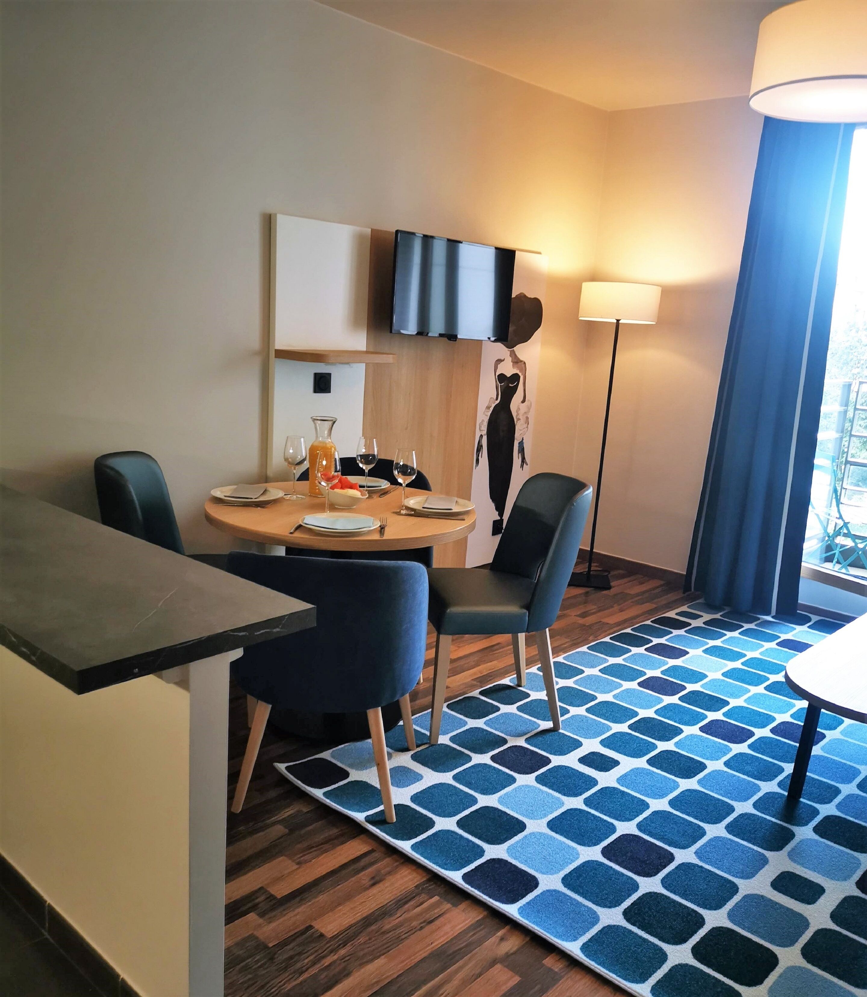 A hotel room with a dining area featuring a round table, two chairs, a TV mounted on the wall, a floor lamp, and a patterned rug.
