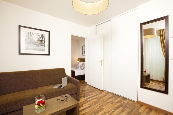 A hotel room with a wooden floor, a brown sofa, a small table with a laptop and a glass of water, a framed picture on the wall, and a mirror reflecting a bed and curtains.