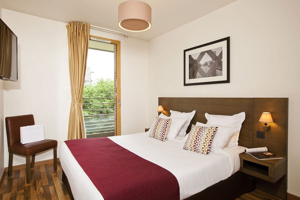 Hotelkamer met een bed, een stoel, een nachtkastje, een lamp, een raam met gordijnen, en een lijstje aan de muur.