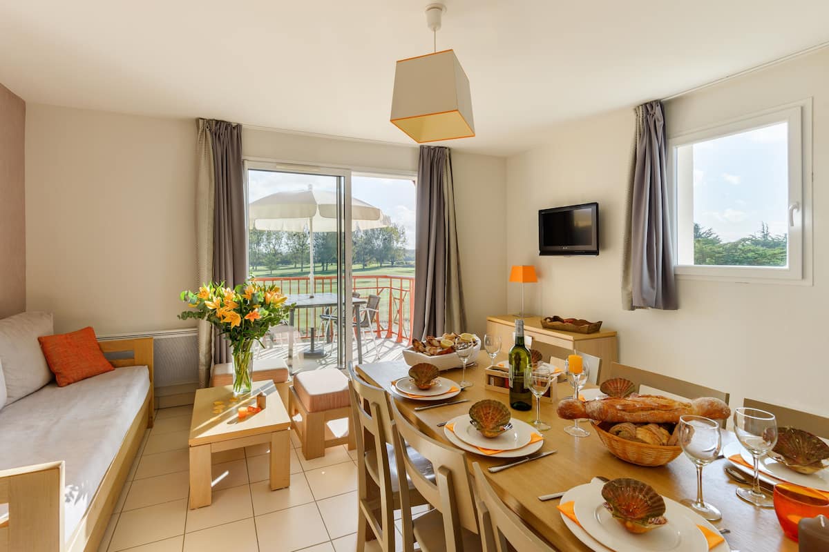 A well-lit room with a dining table set for two, a sofa, a TV, and a view of the outdoors.