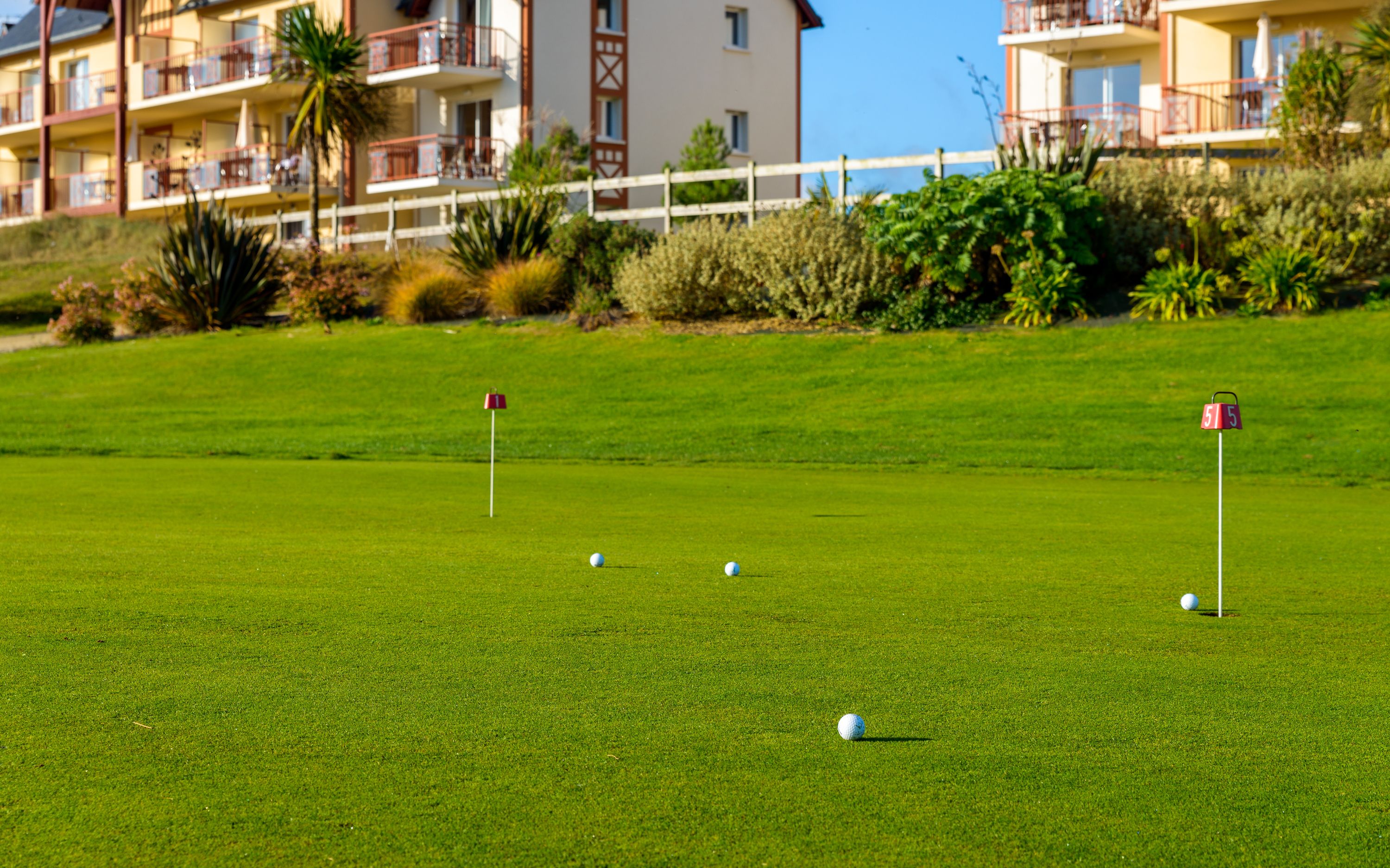 A golf course with a flagstick and golf balls, surrounded by a well-maintained lawn and a building with balconies in the background.