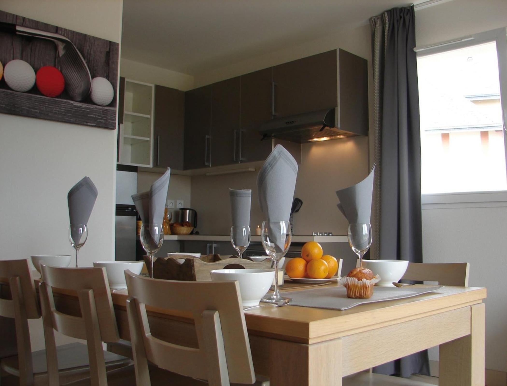 A dining area with a wooden table set for four, glasses, a bowl of oranges, and a muffin on the table.