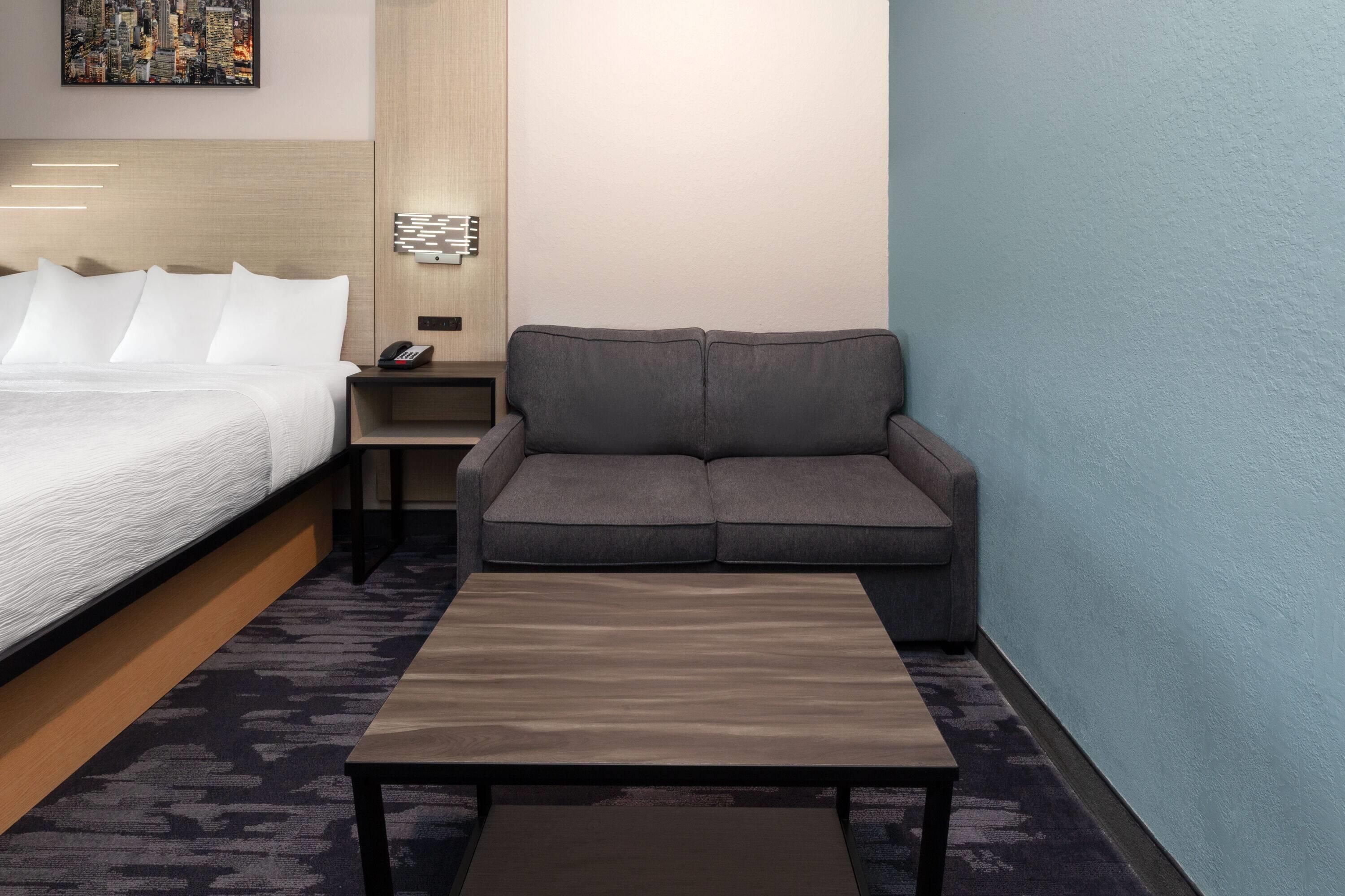A hotel room with a bed, a gray sofa, a small table, and a wall-mounted phone.