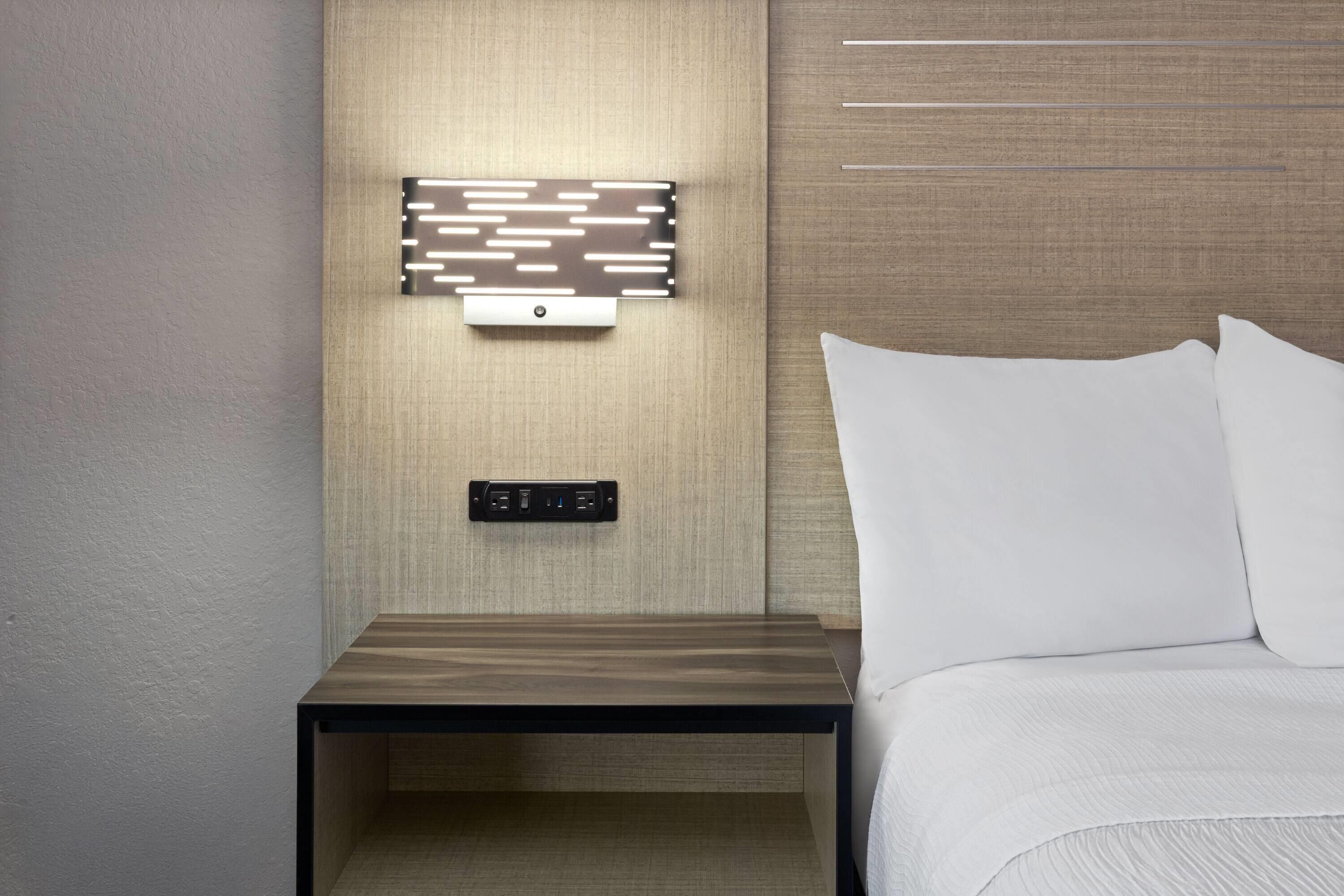 A hotel room with a wooden nightstand, a wall-mounted power outlet, and a bed with white bedding.
