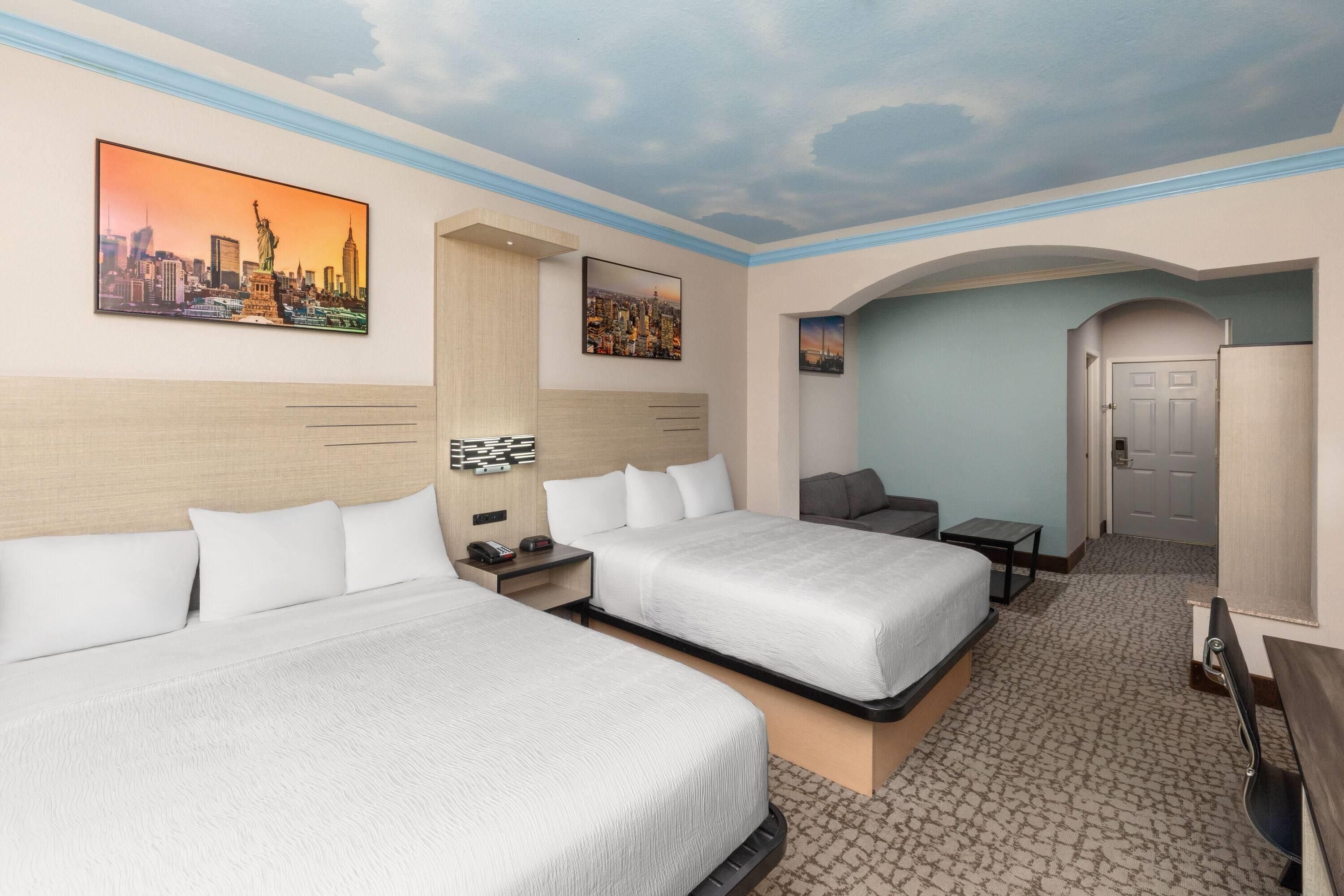A hotel room with two beds, a desk, a chair, and a sofa. There are paintings on the walls and a door leading to another room.