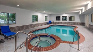 An indoor swimming pool with a brick edge, blue chairs, and a view of the outdoors through windows. - Baymont by Wyndham Texas City (Texas City)