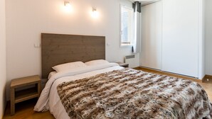 A bedroom with a wooden headboard, a large fluffy blanket, a bedside table, and a window with curtains. - Résidence Lagrange Vacances le Belvédère (Saint-Mamet)