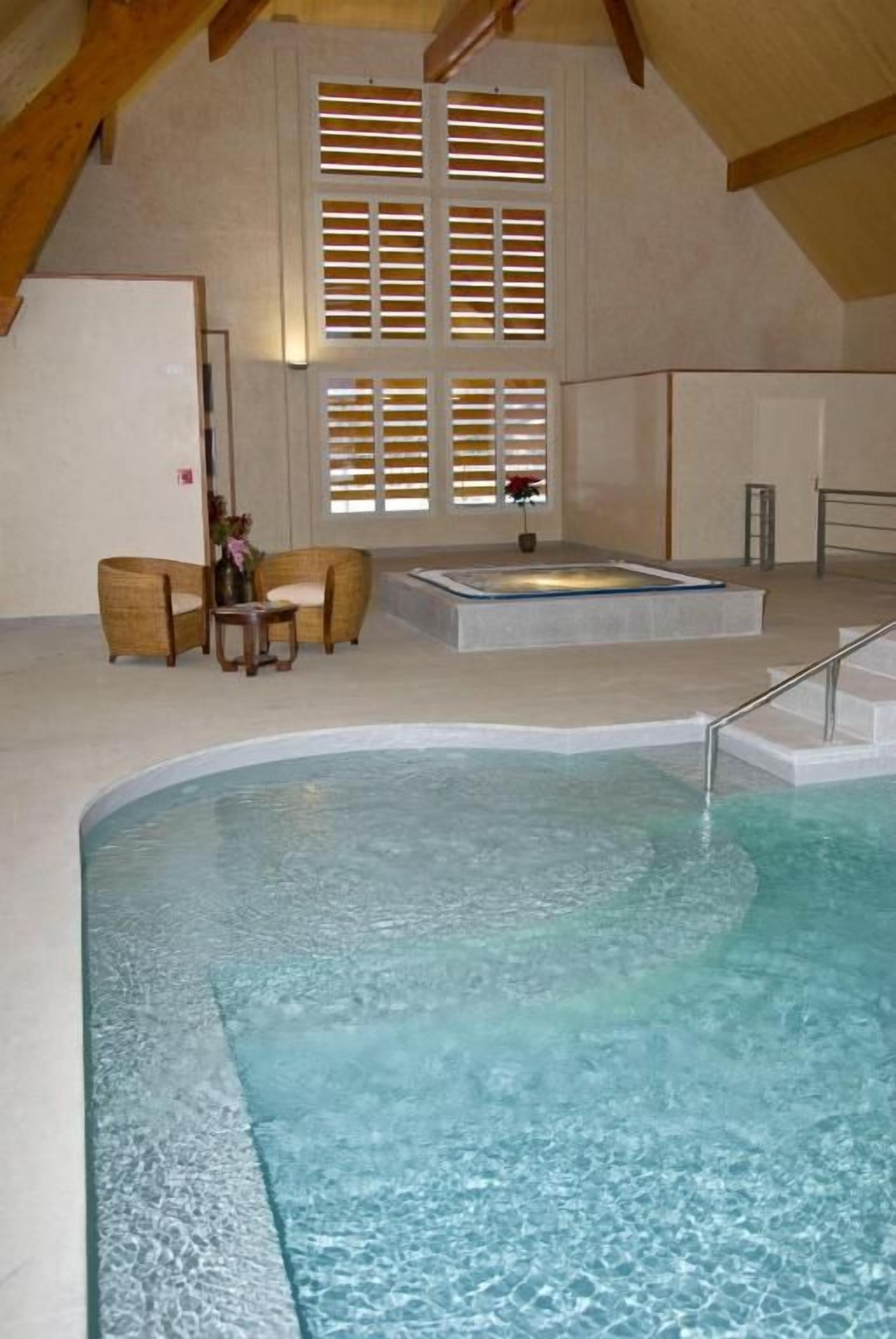 A swimming pool with a jacuzzi, wooden ceiling beams, and a window with blinds.
