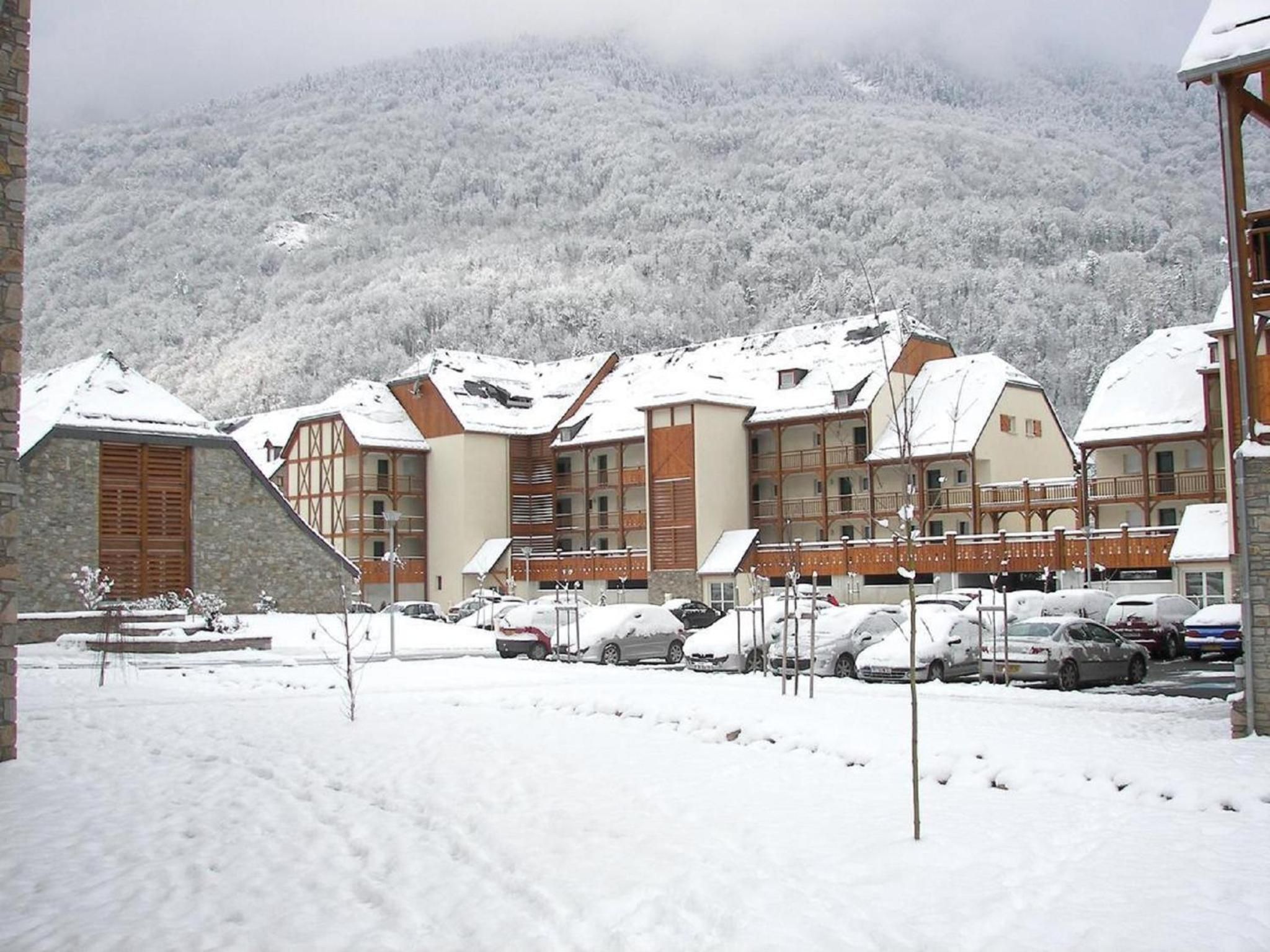 Un edificio cubierto de nieve con varios balcones, un estacionamiento con coches y un paisaje montañoso al fondo.