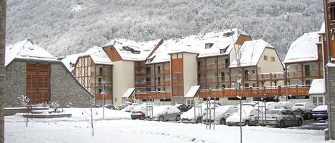 A snow-covered building with multiple balconies, a parking lot with cars, and a mountainous backdrop.