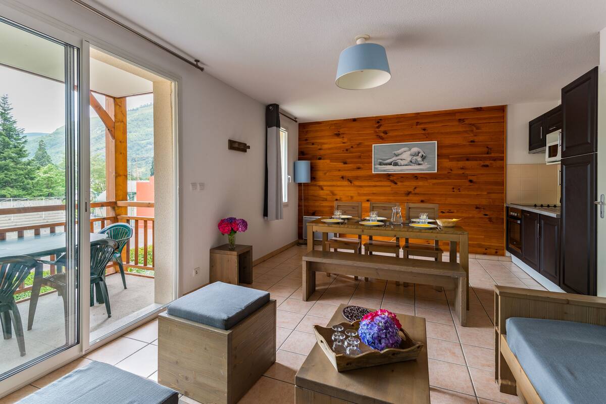 A modern kitchen with wooden accents, a dining area with a table and chairs, and a balcony with a view of greenery.