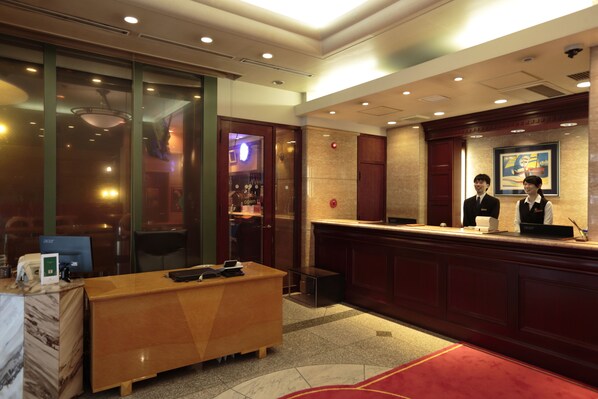 Un espace d’accueil d’hôtel avec un comptoir de réception en bois, un ordinateur et un bar visible à travers les portes vitrées.