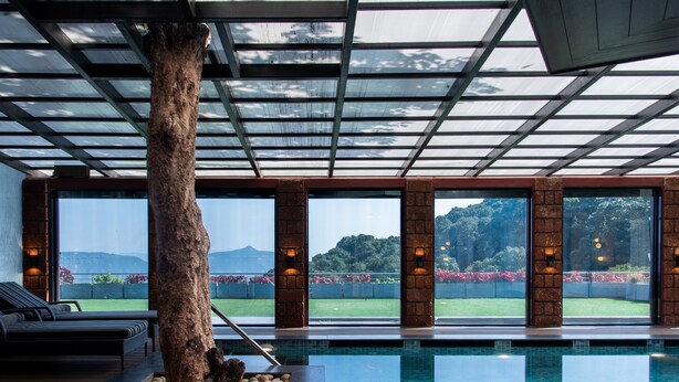 Una piscina interna con un grande tronco d'albero al centro, ampie finestre che offrono vista su un campo da golf e montagne, e una zona relax con vista sulla piscina.