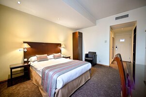 Een hotelkamer met een groot bed, nachtkastjes, een stoel, een kledingkast en een deur die leidt naar een andere kamer.
