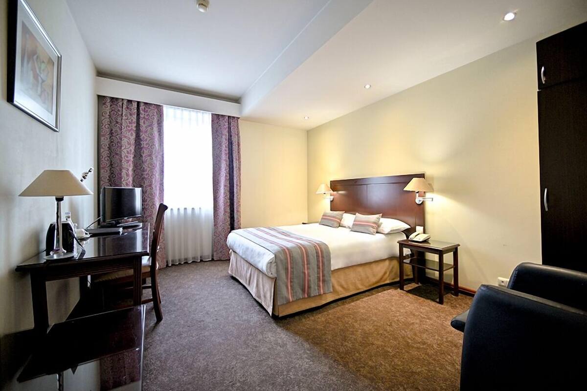 A hotel room with a bed, desk, chair, TV, and lamps.