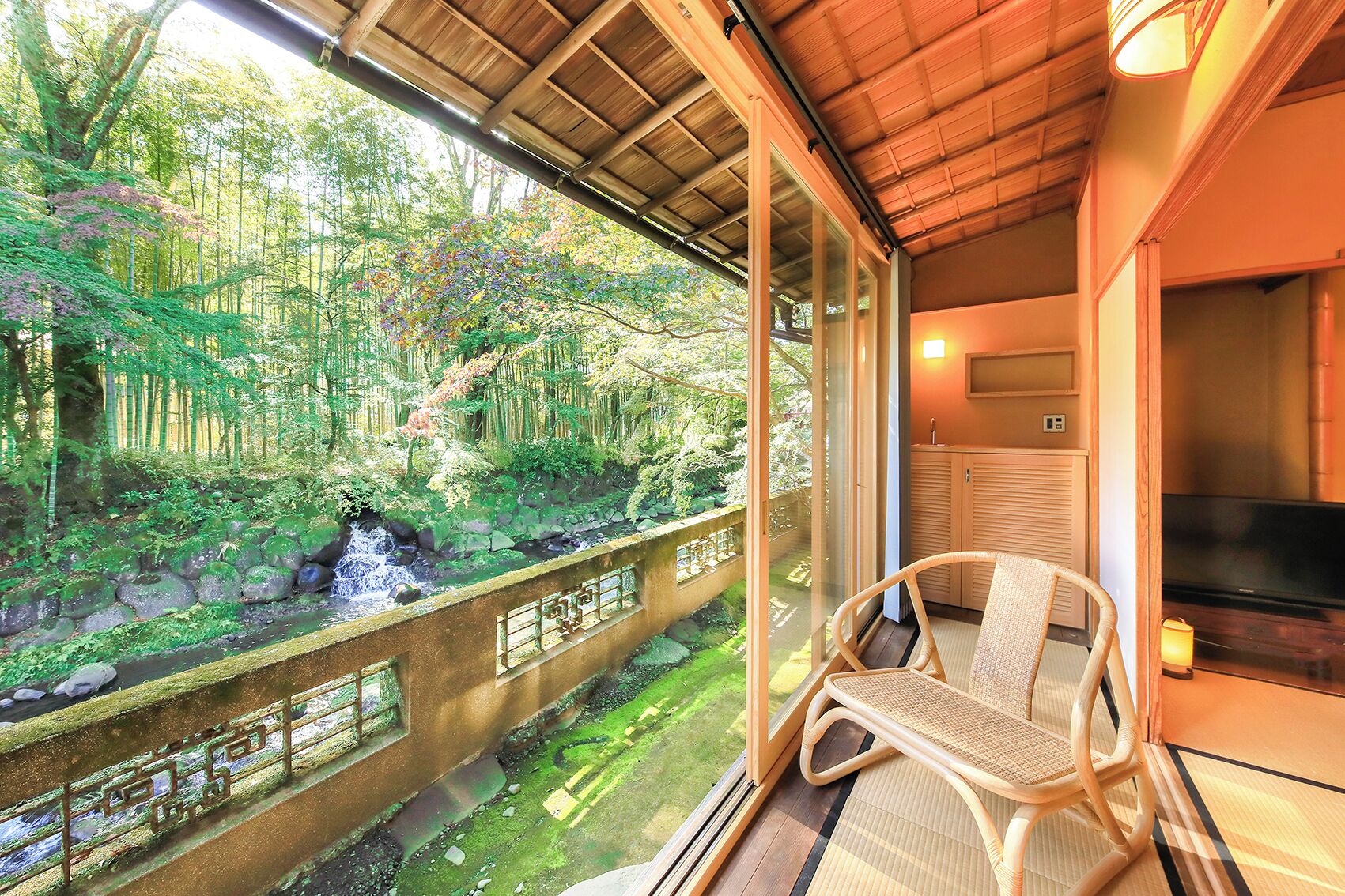 A balcony with a wicker chair, a TV, and a view of a forested area with a stream.