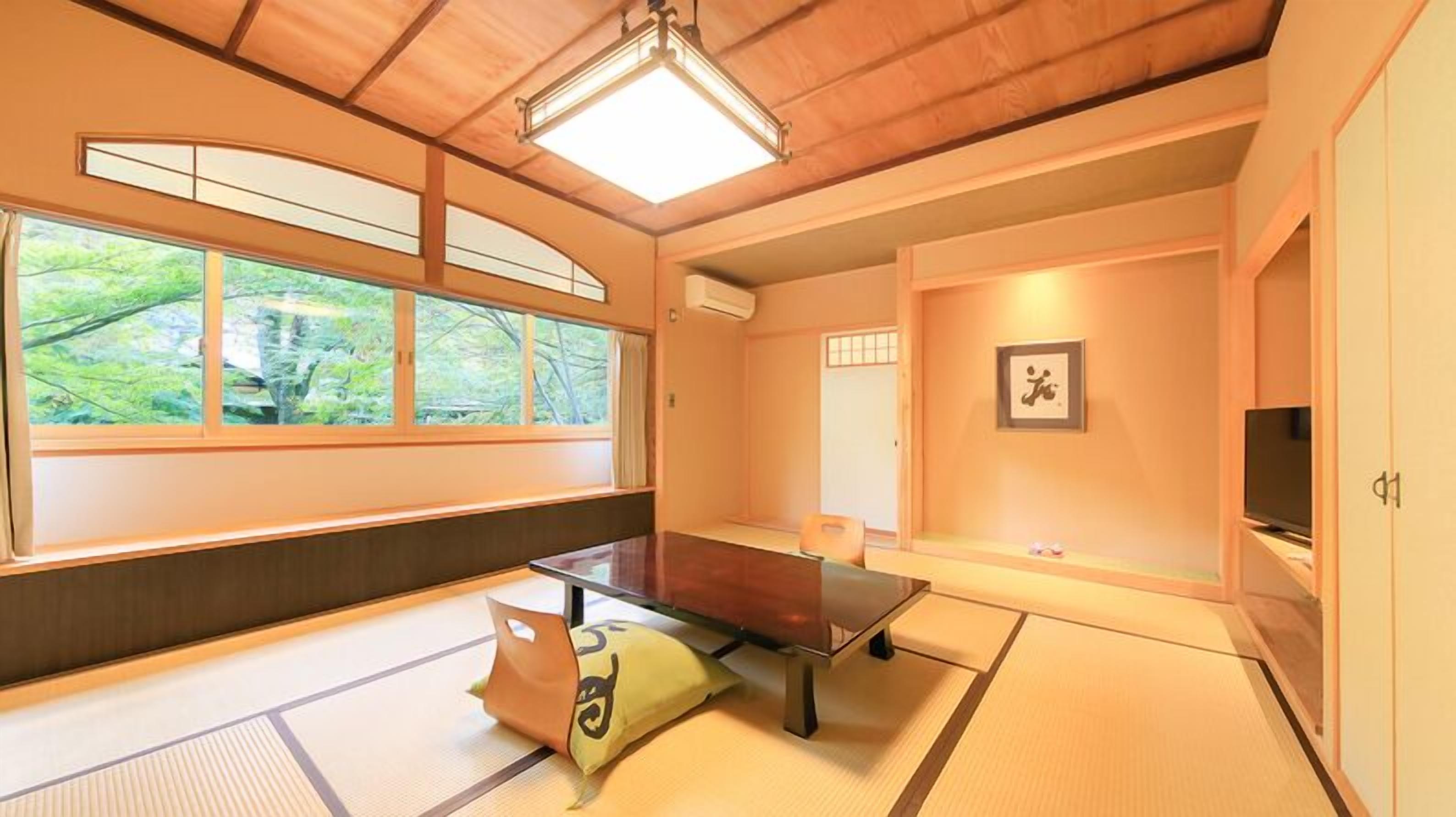 A tatami mat room with a tatami mat table, a futon bed, a television, and a large window with a view of greenery.
