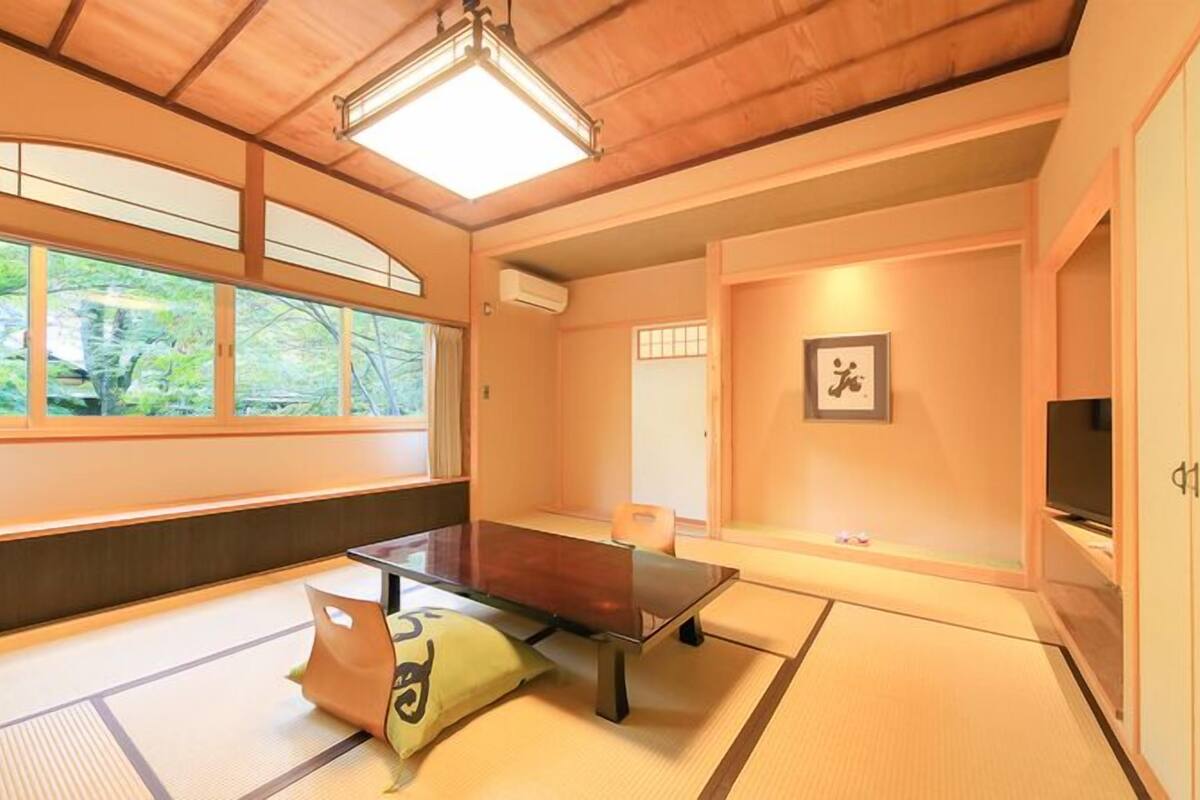 Ein Raum mit Tatami-Matten, einem Tatami-Tisch, einem Futonbett, einem Fernseher und einem großen Fenster mit Blick auf Grünflächen.