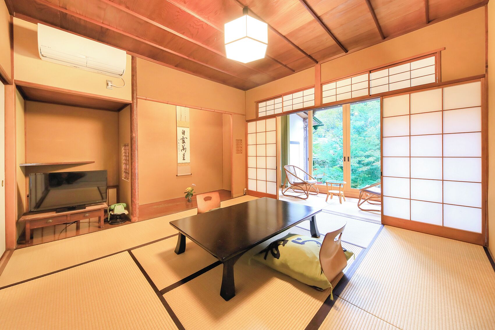 Une pièce de style japonais traditionnel, avec un sol recouvert de tatamis, une table basse, un téléviseur et des portes coulissantes donnant sur un balcon avec vue sur la verdure.