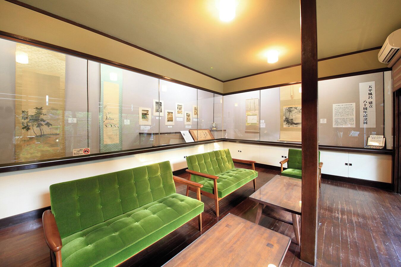 A waiting area with green upholstered chairs, a wooden coffee table, and glass display walls with framed pictures.