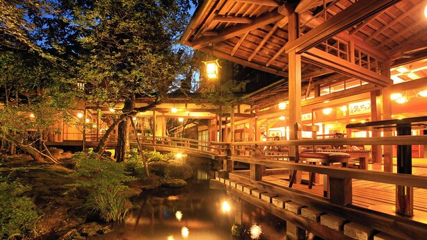 A traditional Japanese-style building with a wooden bridge over a pond, surrounded by trees and illuminated by warm lighting. - Arai Ryokan (Izu)