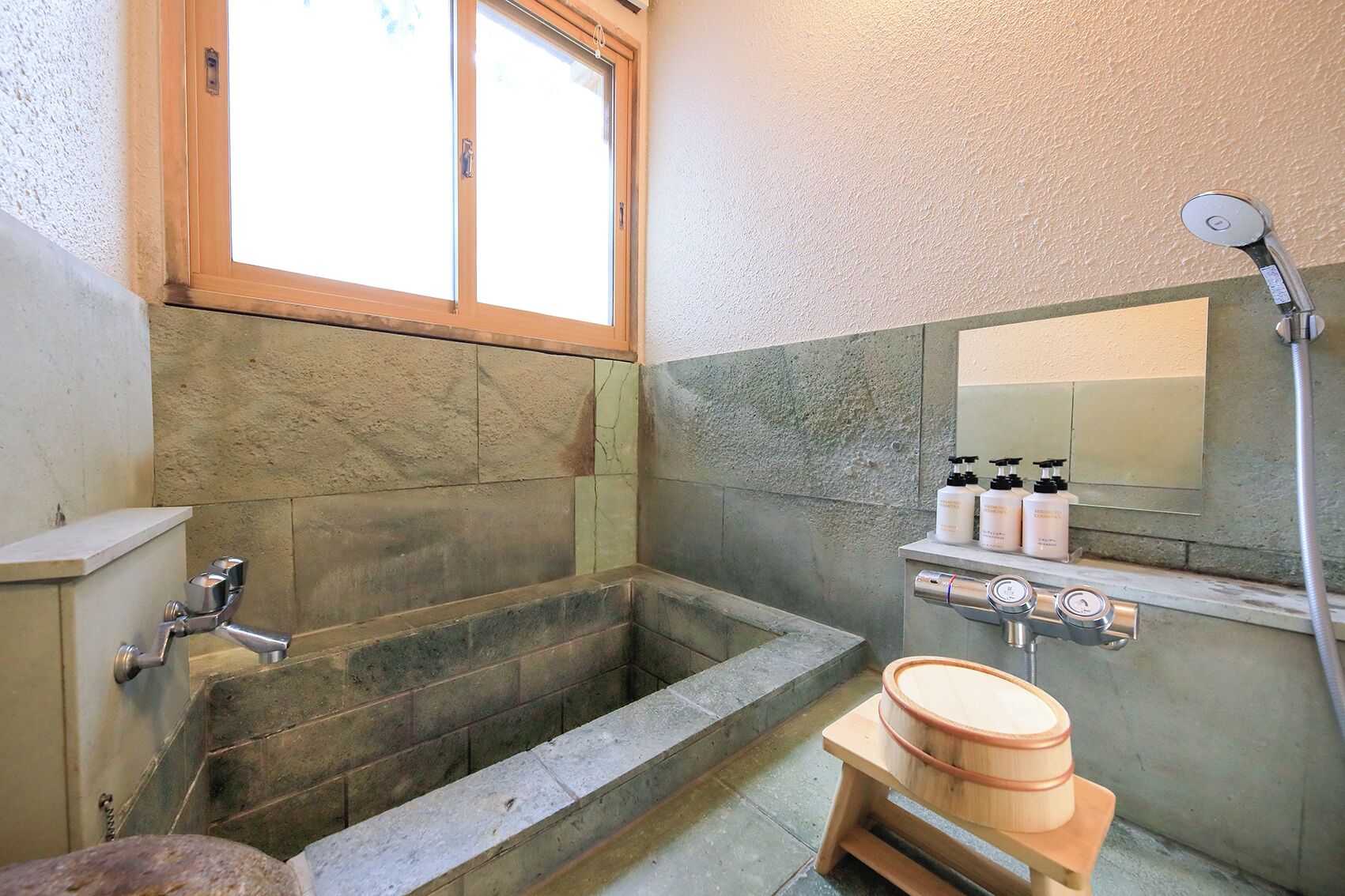 A bathroom with a stone bathtub, a wooden stool, a shower, and a shelf with toiletries.
