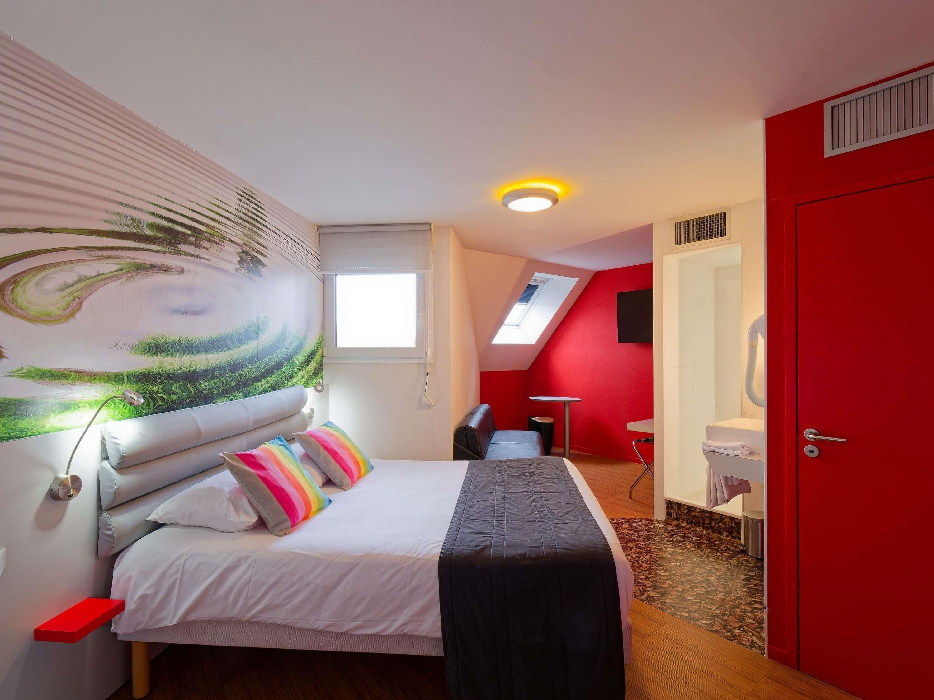 A bedroom with a bed, a red door, a skylight, a small table, and a bathroom with a toilet and sink.