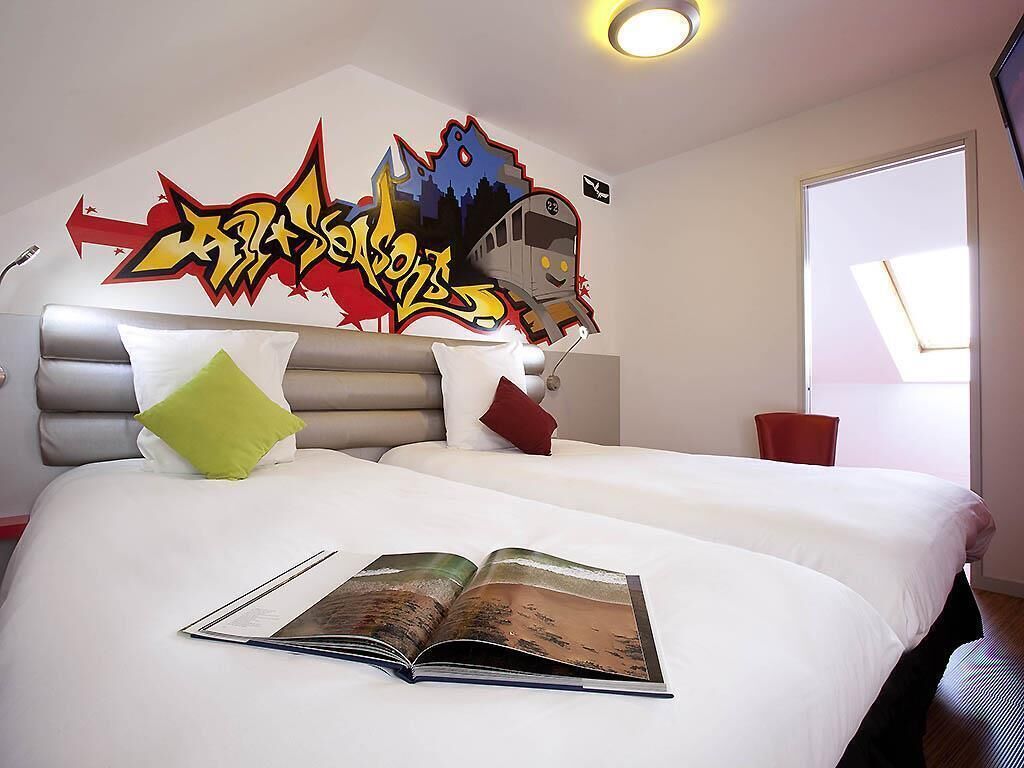 Una habitació d'hotel amb un llit, una gran obra d'art mural amb grafitis, una finestra i una revista damunt el llit.