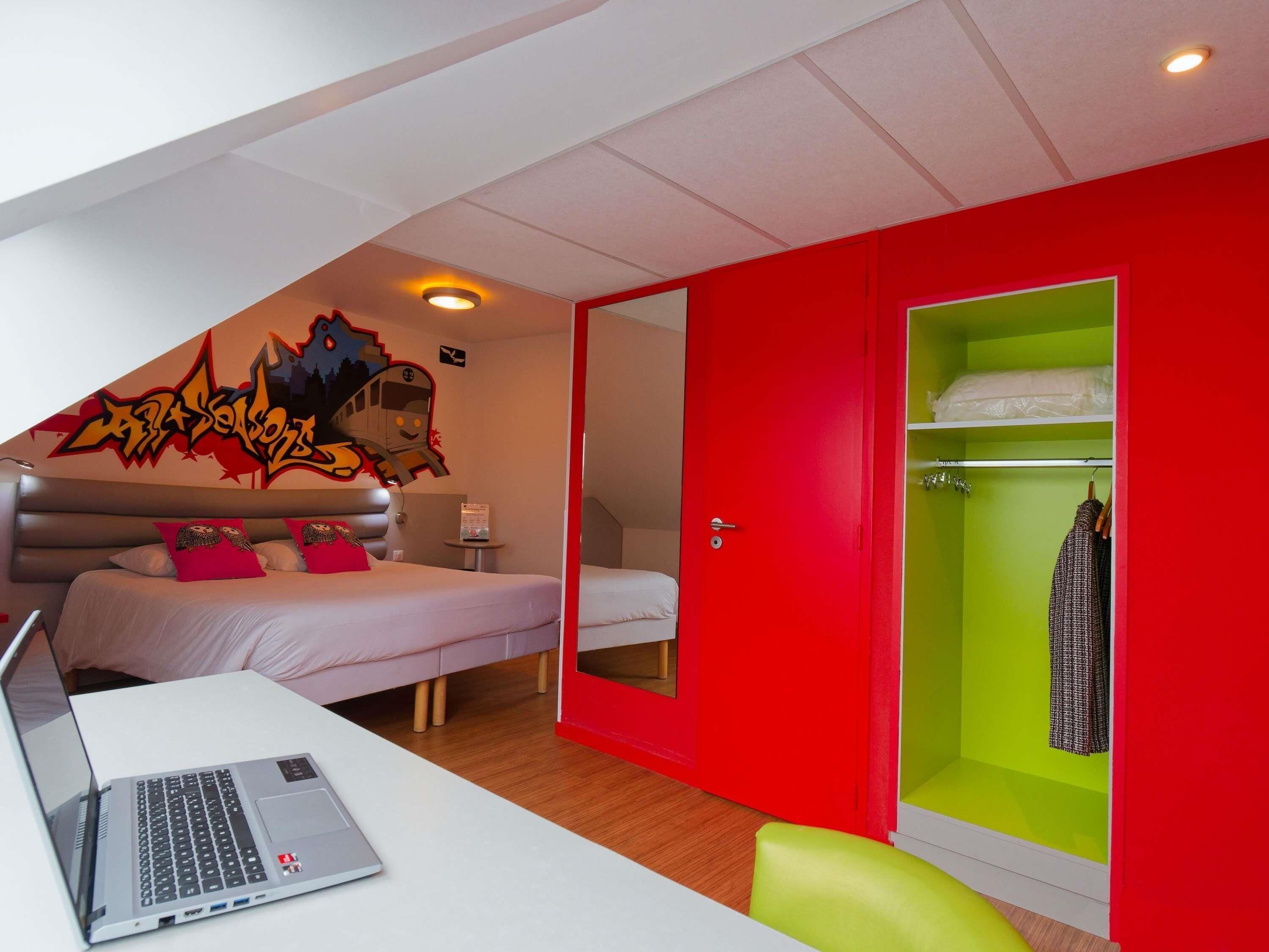 A bedroom with a bed, a desk with a laptop, a red wardrobe, and a green closet.