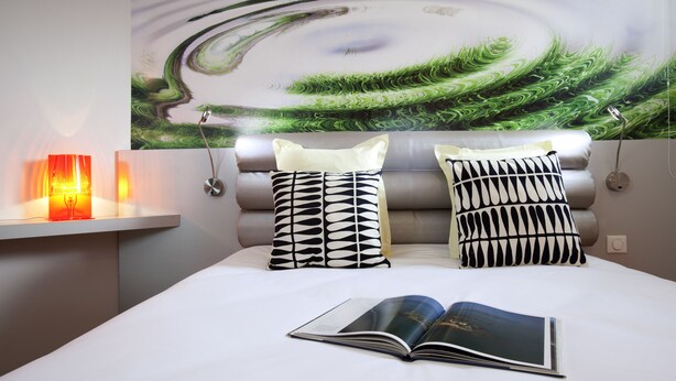 Quarto com cama, duas almofadas estampadas, mesa de cabeceira com abajur e uma grande pintura abstrata na parede.