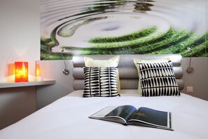 Un dormitorio con una cama, dos almohadas estampadas, una mesita de noche con una lámpara y un cuadro abstracto grande en la pared.