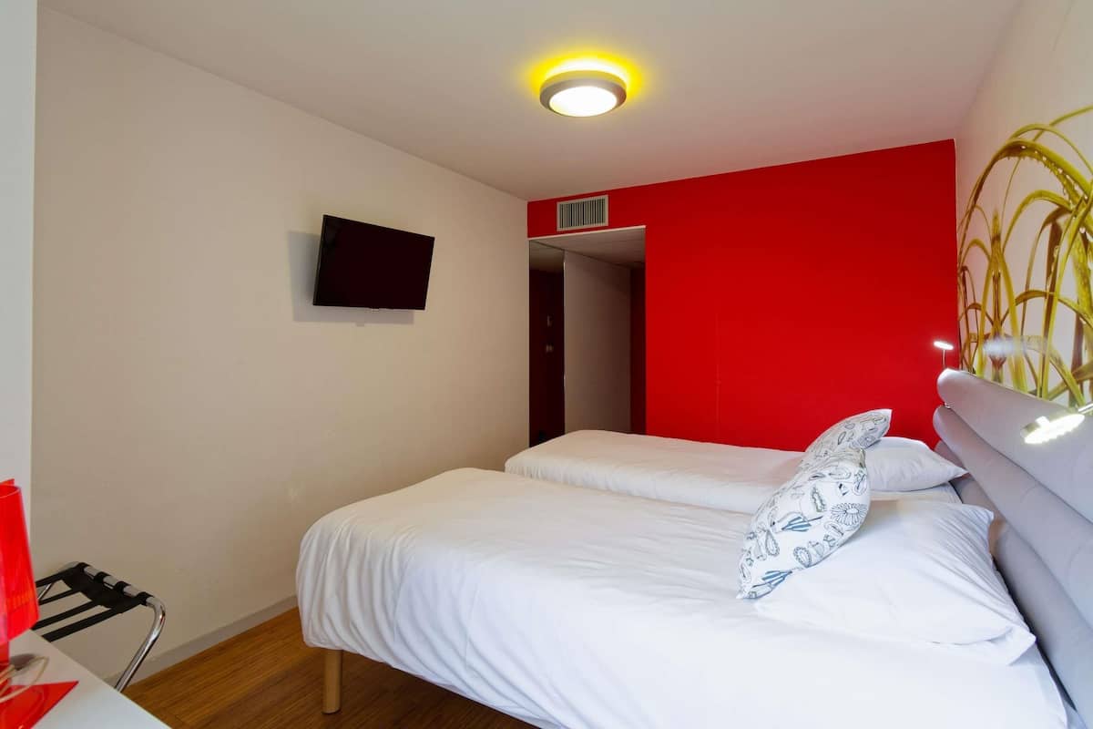 A hotel room with a bed, a TV, a red accent wall, and a ceiling light.