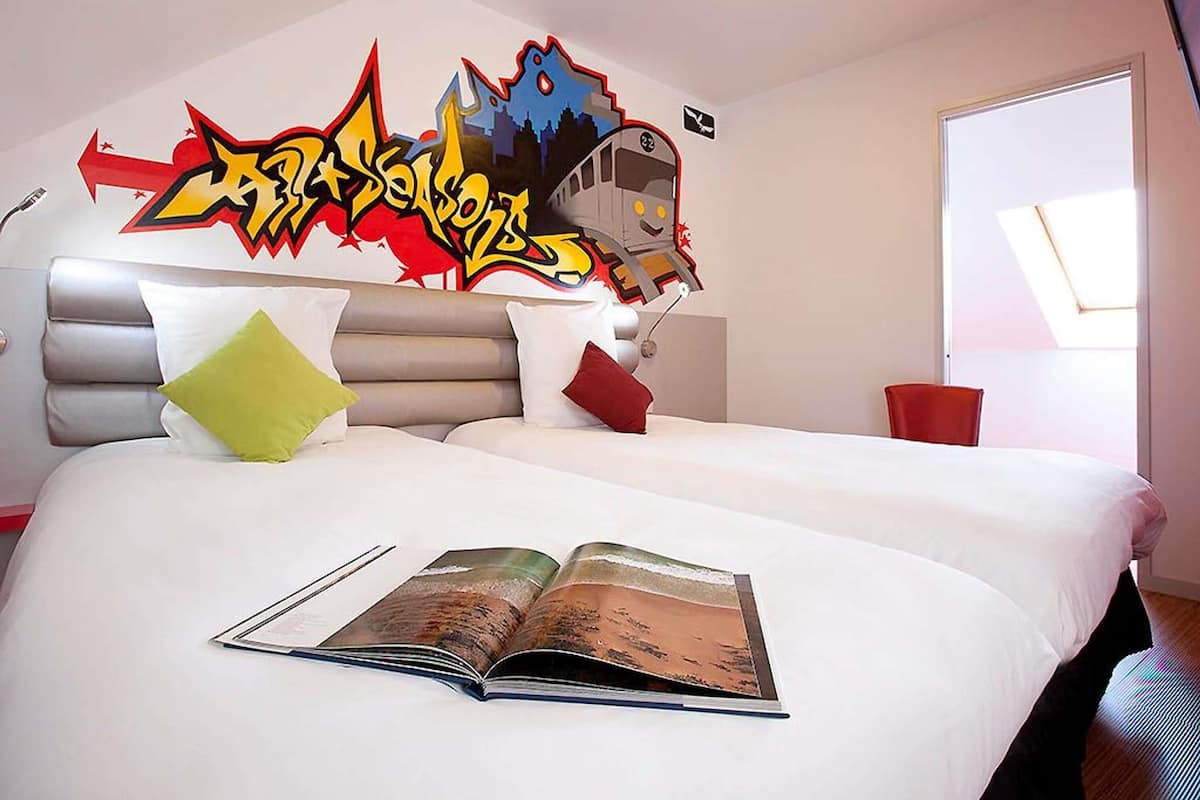 A modern hotel room with a large bed, a graffiti wall art, a window with a view, and a book on the bed.
