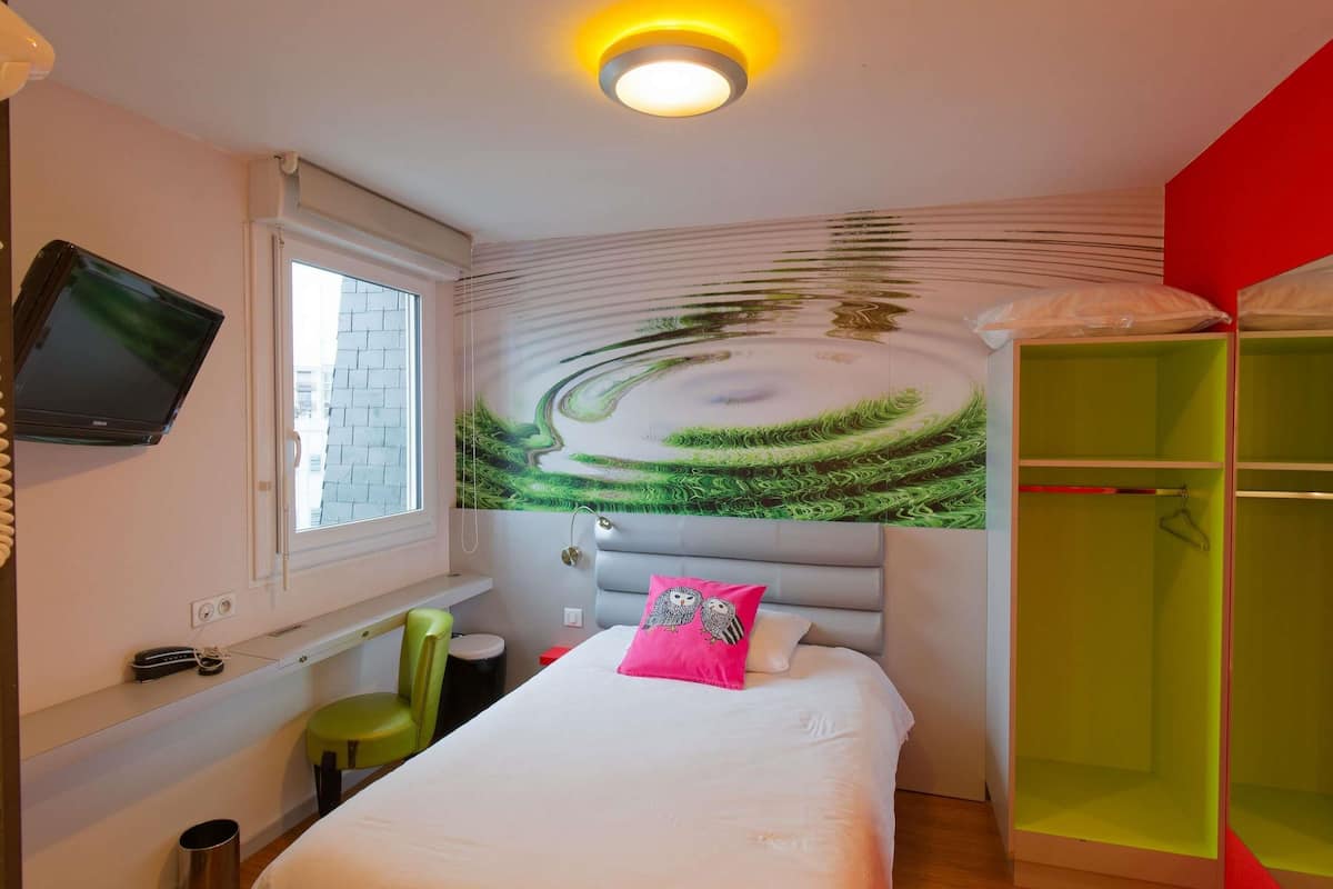 A hotel room with a bed, a TV, a window, a green chair, a red wardrobe, and a wall mural.