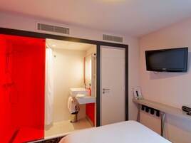 Habitación de hotel con ducha, lavamanos, inodoro, cama, televisor y un pequeño estante con un teléfono y un control remoto.