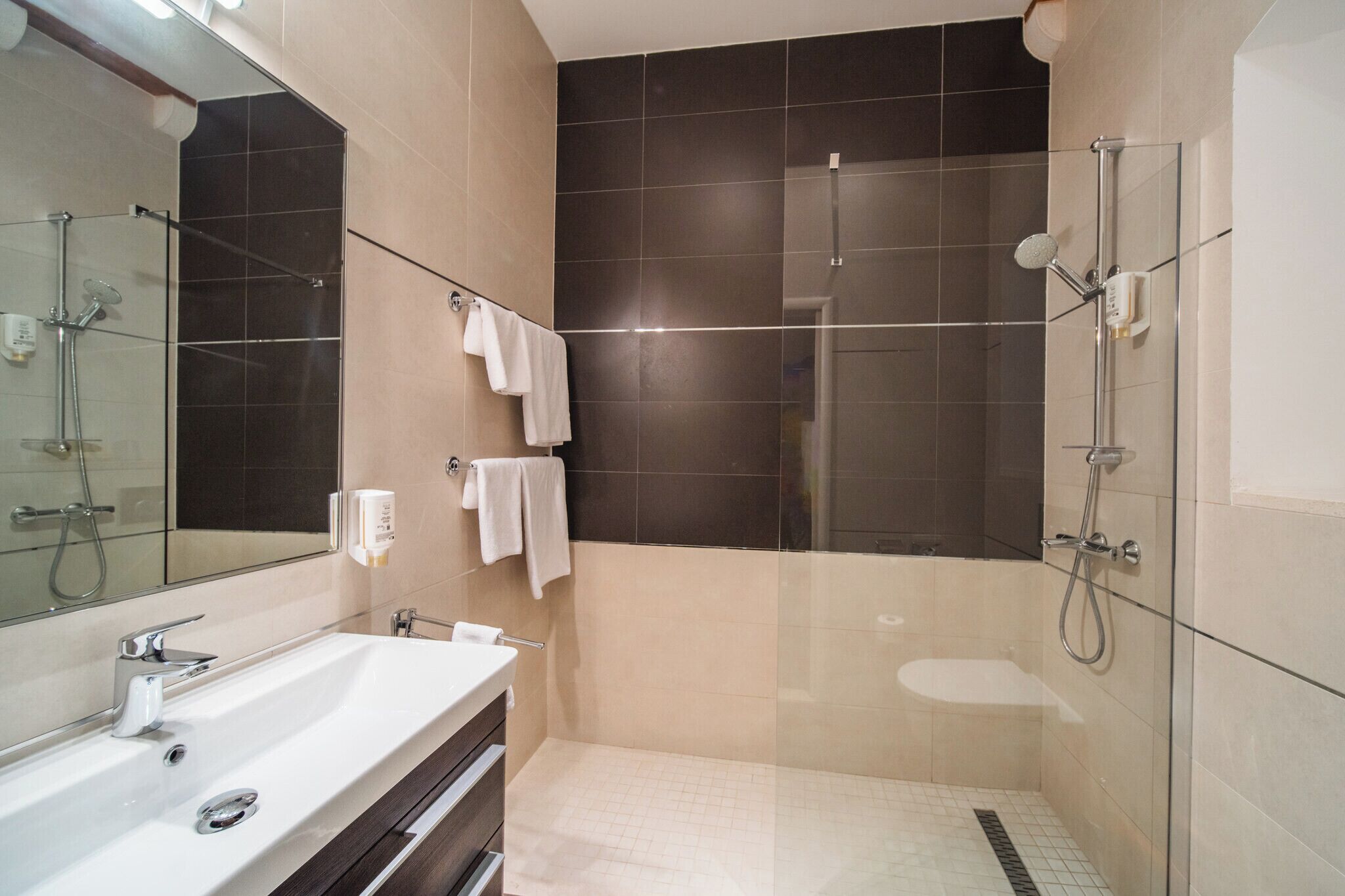 Ein modernes Badezimmer mit einem großen Spiegel, einem weißen Waschbecken, einer Duschkabine aus Glas und zwei weißen Handtüchern an der Wand.