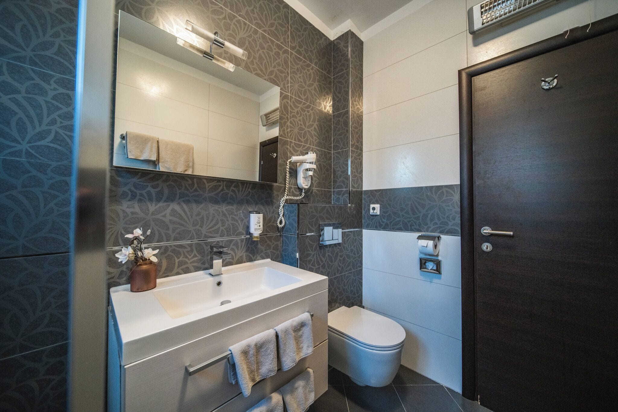A modern bathroom with a large mirror, a white sink, a towel rack, a toilet, and a dark wooden door.