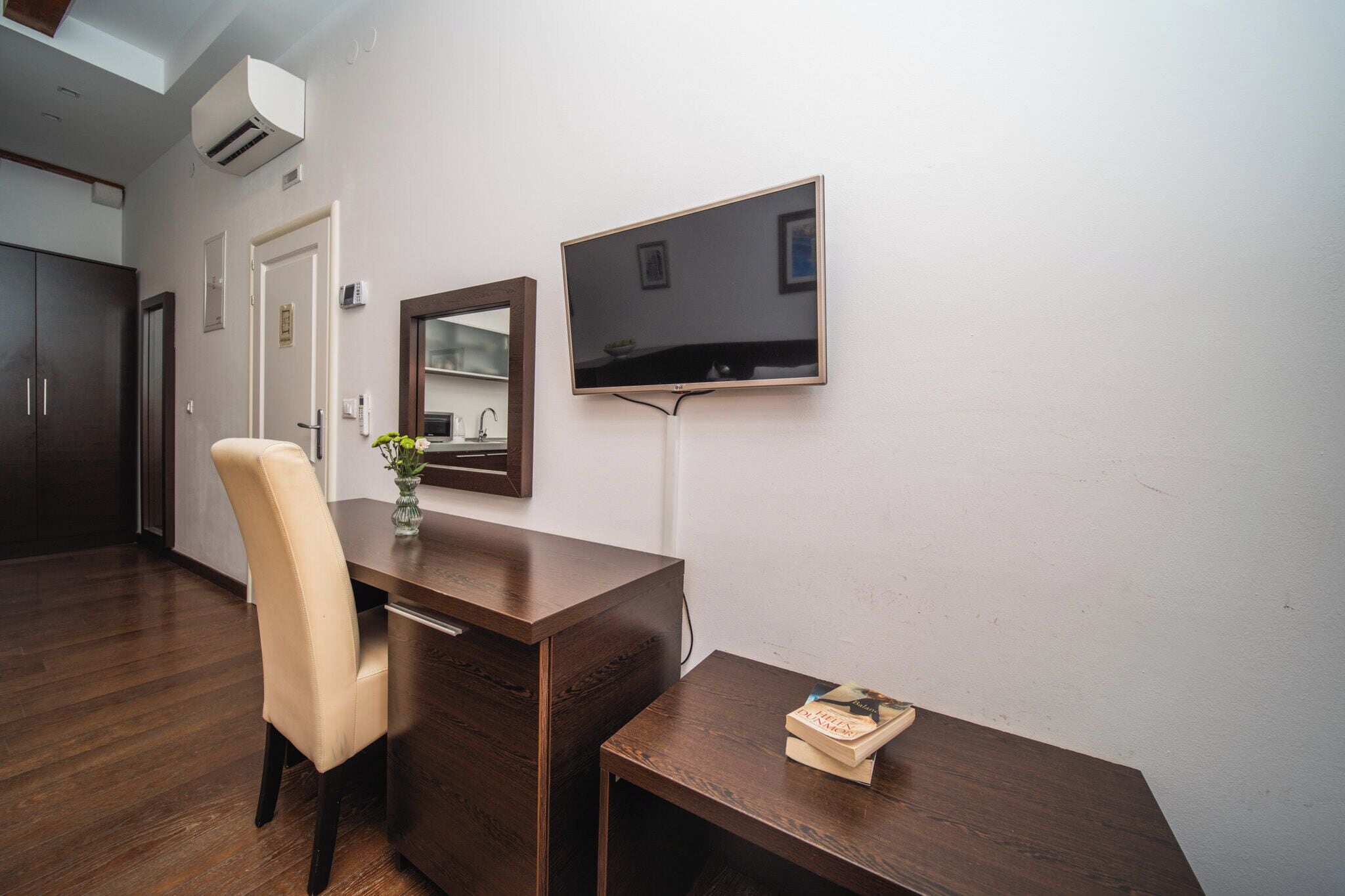 Una habitación moderna con un escritorio de madera, una silla, una pequeña mesa con libros, un televisor de pantalla plana montado en la pared y un espejo sobre el escritorio.