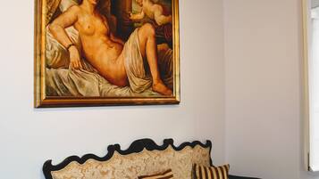 A room with a large classical painting on the wall, a decorative sofa with patterned cushions, and a window with a white frame.