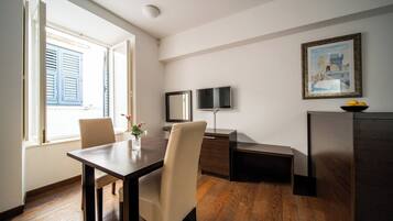 A room with a wooden dining table, two chairs, a TV, a small cabinet, and a painting on the wall.