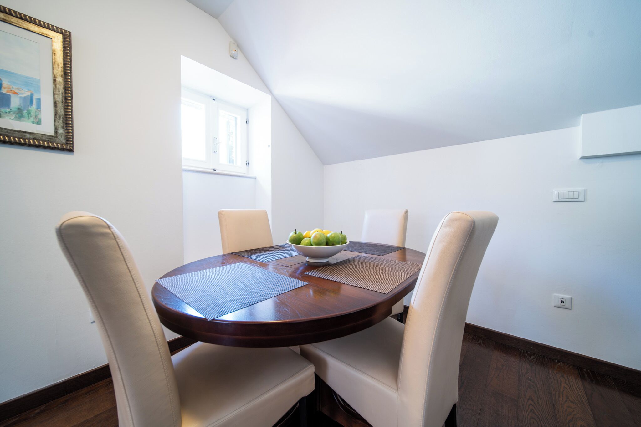 Un coin repas avec une table ronde, quatre chaises, un saladier de fruits, une fenêtre et un tableau encadré au mur.
