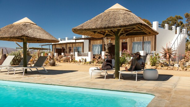 A poolside area with lounge chairs and thatched umbrellas, a pool, and a building with a white facade and wooden accents. - Inverdoorn Private Game Reserve (Breede River)