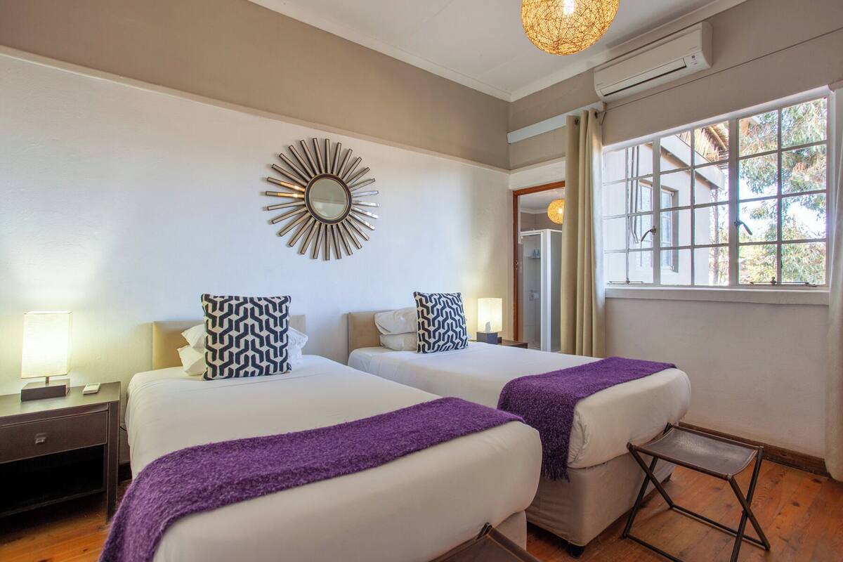 A hotel room with two beds, a sunburst mirror, a window with a view, and a small table.