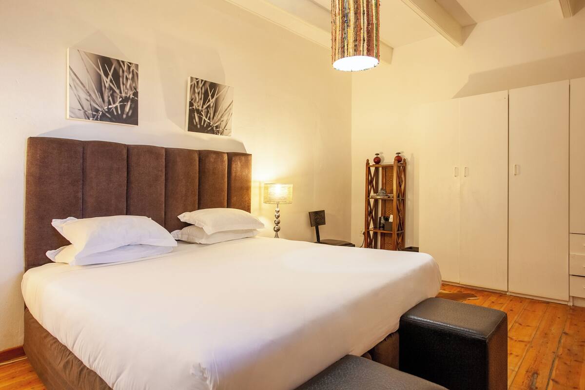 A neatly made bed with white linens, a wooden headboard, and a bedside lamp. There is a wooden bench at the foot of the bed, and a wardrobe with a mirror.