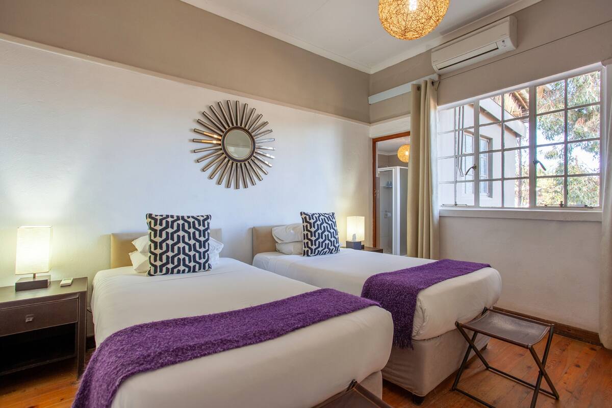 A hotel room with two beds, a mirror, a window with curtains, and a small table.